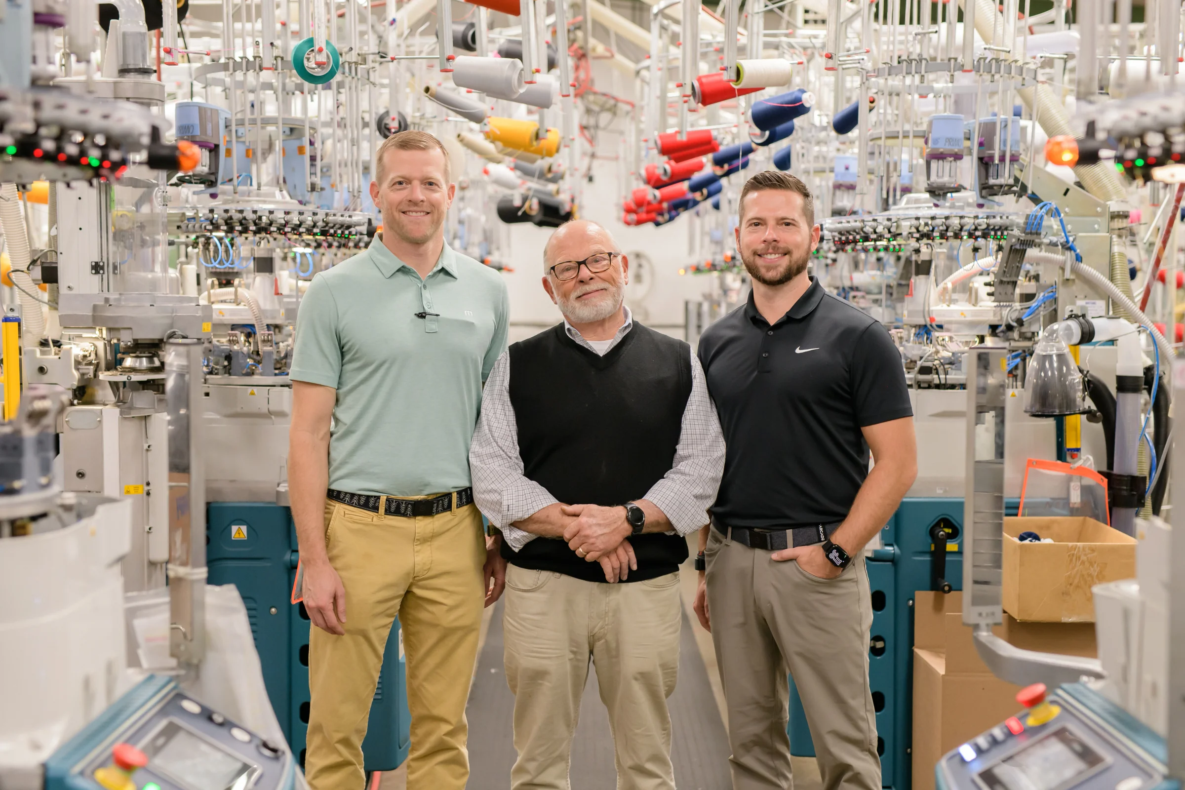 Three men standing among knitting machines inside the sock mill